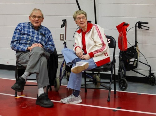 Sue sits wearing a white and red jacket. A man sits beside her.