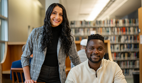 A young woman stands and smiles next to a young man who is sitting down and smiling.