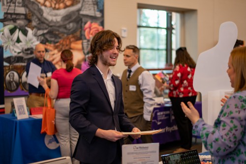 A young man wears a suit and smiles while listening to a woman explain something.