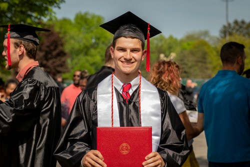 A young man smiles while holding his diploma after commencement.