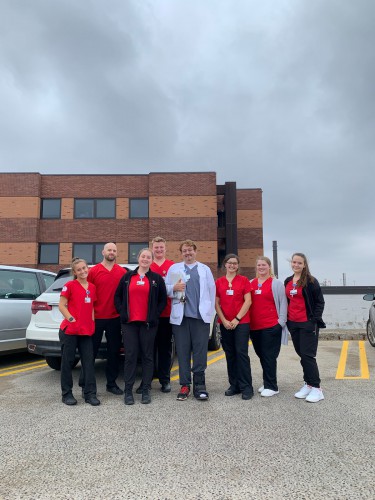 8 people wear nursing or medical attire and stand in a parking lot, smiling.