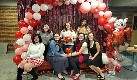 A group of 7 young woman are in front of a red, pink, and white balloon arch. 4 sit on a bench and 3 stand behind them.