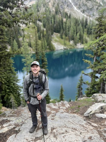 Jason stands on mountain with hiking gear. There are water and trees in the background.