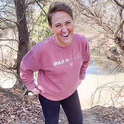 Kristey wears a pink sweater and smiles in front of a tree and body of water.