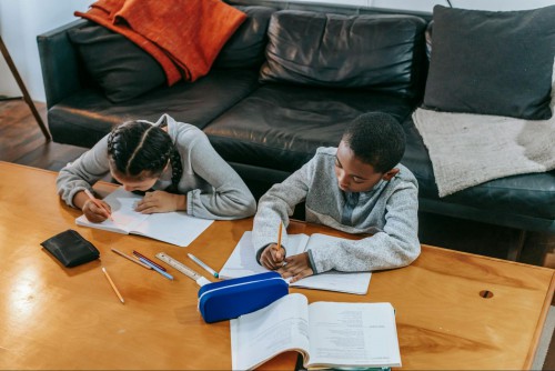 2 children work on schoolwork while sitting on the floor leaning against a couch. A low table is in front of them.