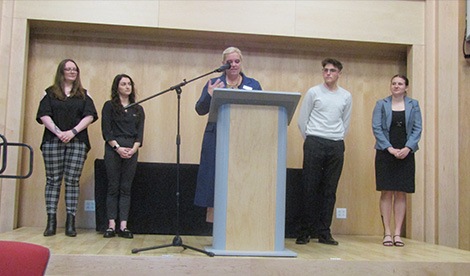 Professor Laura Falco speaks and the winners of the essay competition stand behind her.