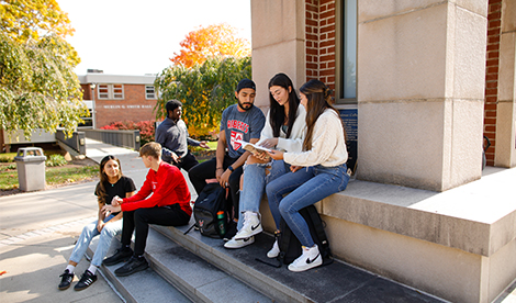 6 students gather around the clock tower on campus. 3 sit and study together. 2 sit on a step below them and talk. The sixth is alone, standing in the background, talking to someone out of the camera shot.