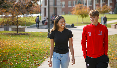 2 students face forward and walk on campus, talking to each other. The clock tower is seen in the background.