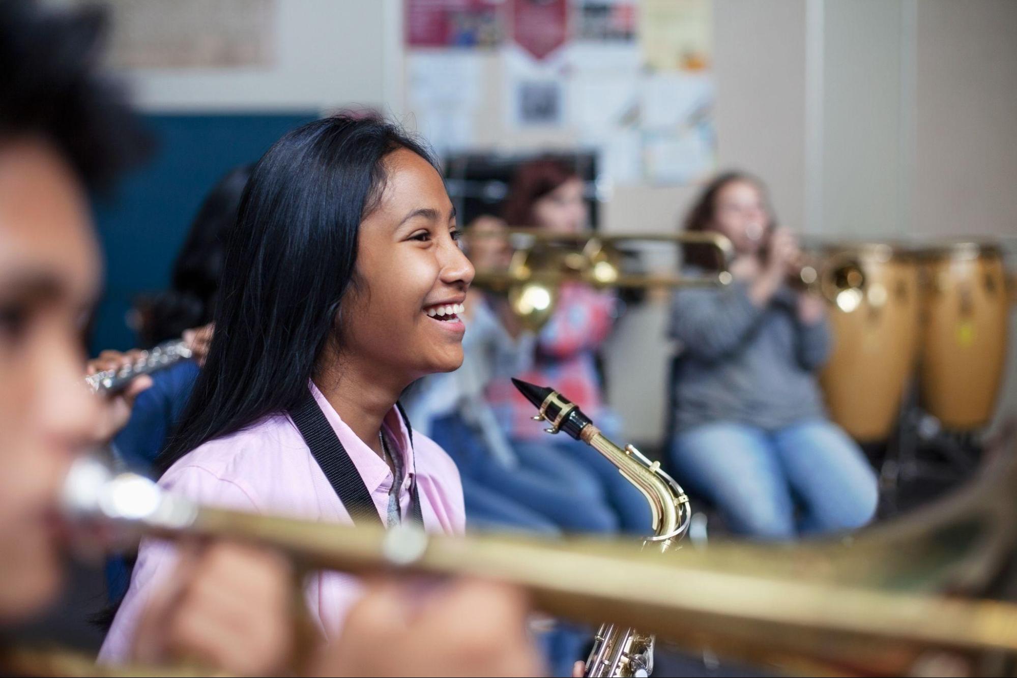 A young woman holds a saxophone and smiles. Other students playing instruments are visible around her.