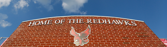 A brick wall that says "Home of the Redhawks" and has the Roberts Redhawk logo. The blue sky is visible above with some white clouds.