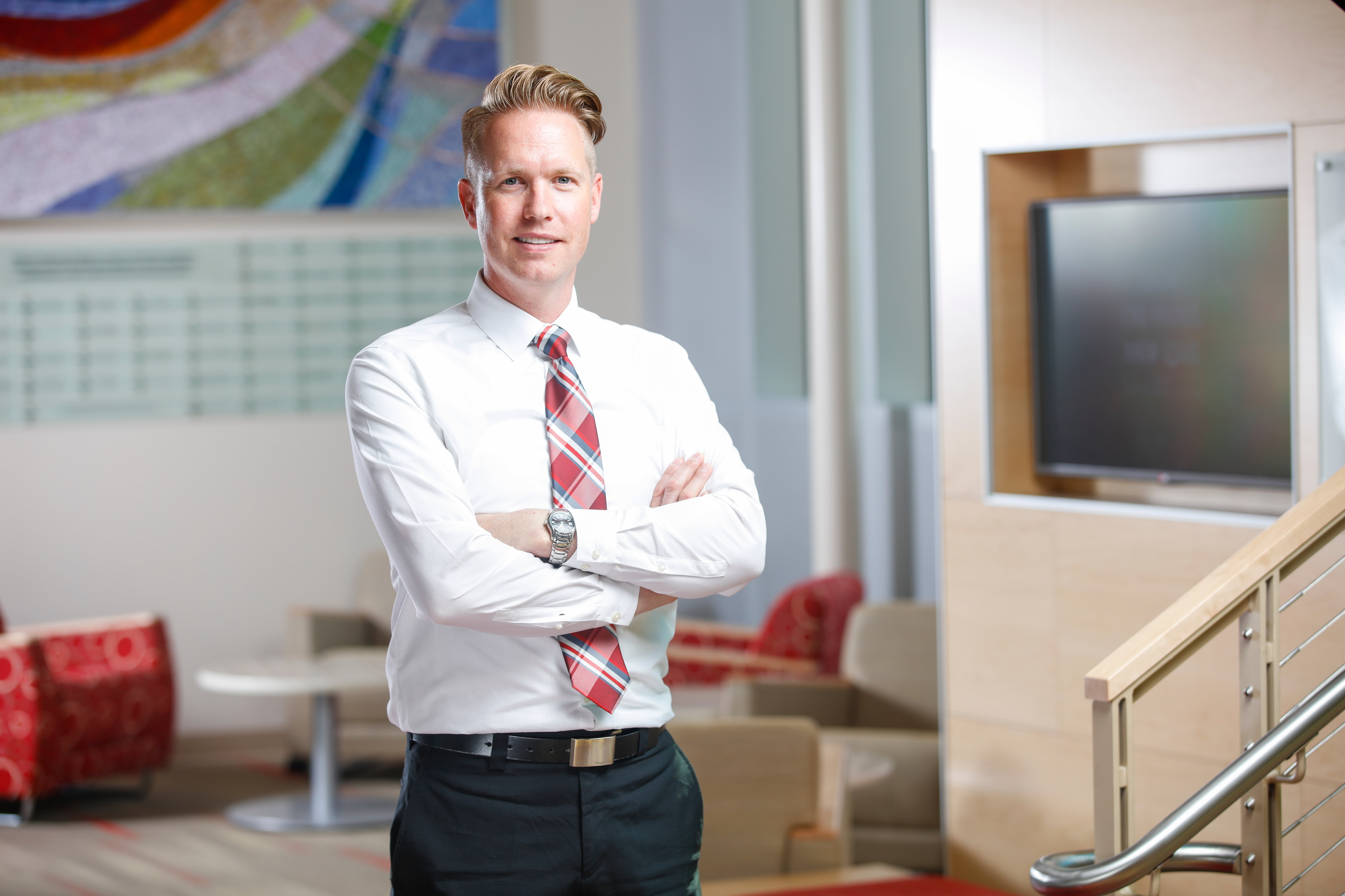 A business man wearing a white dress shirt, red plaid tie, and black trousers stands confidently with his arms crossed